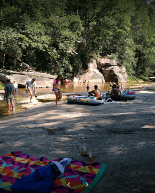People enjoying a sunny day by a river, with some swimming and others relaxing on the rocks and a colorful blanket nearby.