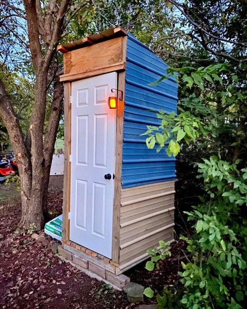 A small, colorful shed with a white door, surrounded by trees and greenery, illuminated by a hanging light.
