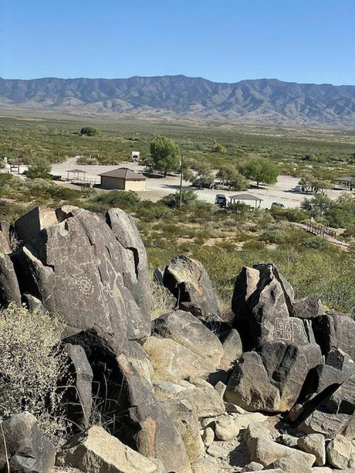 A rocky landscape with petroglyphs, overlooking a valley and distant mountains under a clear blue sky.