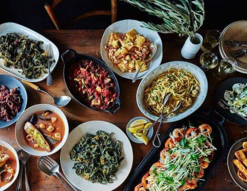A variety of colorful dishes on a wooden table, including pasta, seafood, and greens, with drinks and utensils.