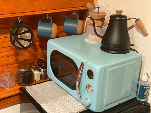 A light blue microwave on a countertop with a coffee maker, mugs, and various kitchen items in a cozy setting.