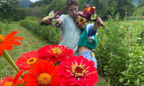 A couple poses playfully in a flower field, holding colorful blooms with vibrant flowers in the foreground.
