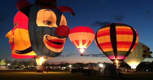 Colorful hot air balloons, including a clown face design, illuminated against a twilight sky.