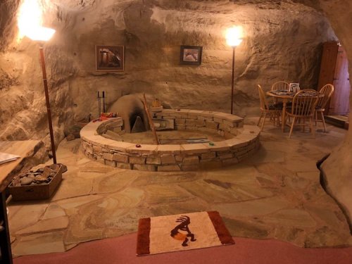 Cozy cave interior with a stone fire pit, wooden furniture, and warm lighting, featuring a decorative rug.
