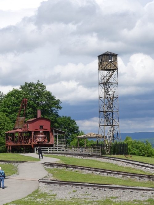 A red vintage train engine near a tall metal tower, set against a backdrop of green trees and cloudy skies.
