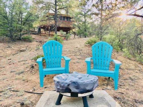 Two blue Adirondack chairs facing a fire pit, surrounded by trees and a cabin in the background. Sunlight filters through.
