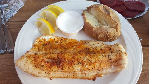 A plate with seasoned fish fillet, lemon wedges, a baked potato, and a small cup of sour cream.