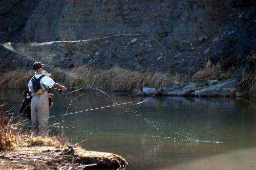 A person in waders casts a fishing line into a calm river surrounded by rocky banks and dry grass.