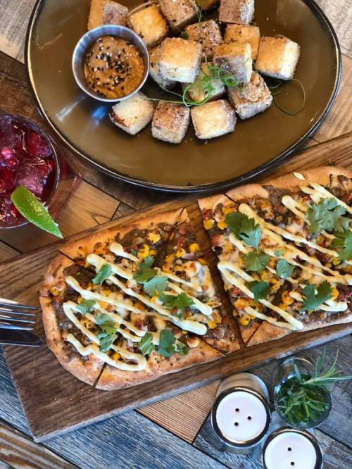 A wooden platter with flatbread pizza topped with corn and cilantro, alongside a plate of crispy cubes and a drink.