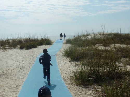A child runs along a blue path on the beach, leading to two people walking towards the ocean under a cloudy sky.
