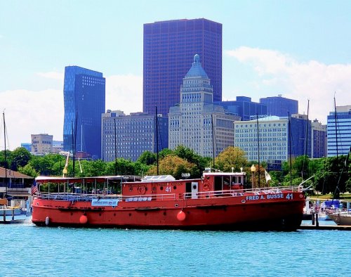 A red boat named "Fred A. Busse" docked in a harbor with a city skyline in the background on a sunny day.