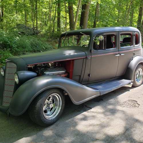 A vintage black car parked on a tree-lined road, showcasing classic design and shiny chrome wheels.