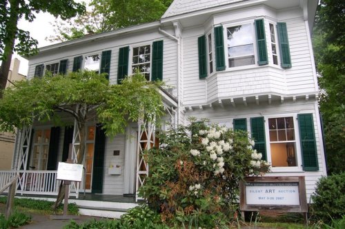A white house with green shutters, surrounded by trees and flowers, featuring a sign for a silent art auction.