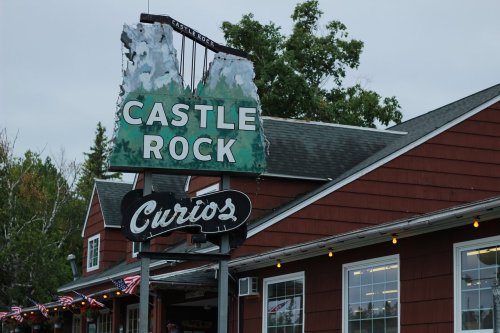 Sign for Castle Rock and Curios, featuring a rustic design and surrounded by trees, with a cloudy sky above.