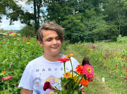 A boy stands among colorful flowers, smiling as a butterfly rests nearby in a lush garden.