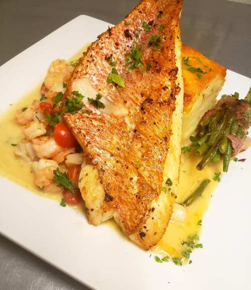 A beautifully plated dish featuring seared fish, shrimp, vegetables, and a side of green beans, garnished with herbs.