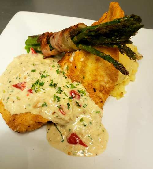 A plate featuring fried fish topped with creamy sauce, served with mashed potatoes and bacon-wrapped asparagus.