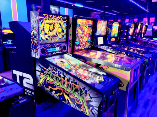 A row of colorful pinball machines in a dimly lit arcade, featuring vibrant artwork and neon lights.