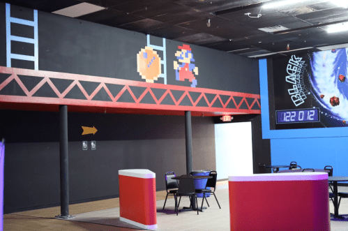 A colorful arcade interior featuring a mural of a pixelated character and a ladder on the wall, with tables and chairs.
