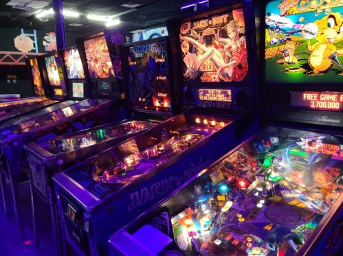 A row of colorful pinball machines with illuminated displays in a dimly lit arcade.