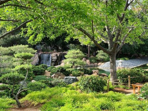 Lush green garden with a waterfall, rocks, and a serene pond surrounded by trees and shrubs.