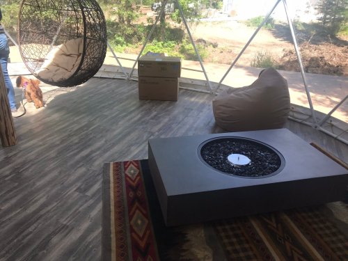 Cozy interior of a geodesic dome with a fire pit, bean bags, and a hanging chair, surrounded by nature.