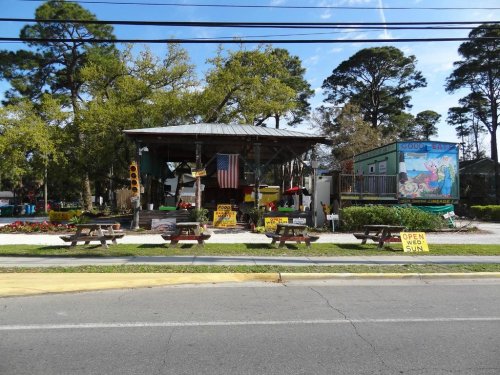 A roadside market with picnic tables, colorful signs, and an American flag, surrounded by trees and a clear blue sky.
