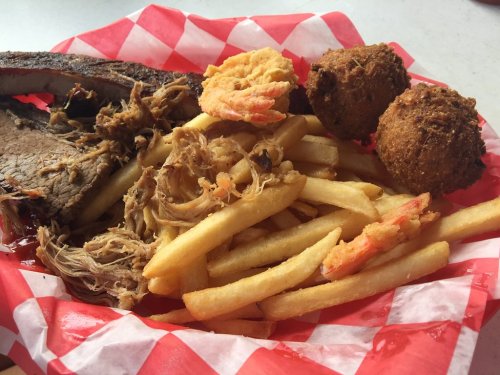 A basket of barbecue featuring ribs, pulled pork, fries, shrimp, and two hushpuppies on a red and white checkered paper.