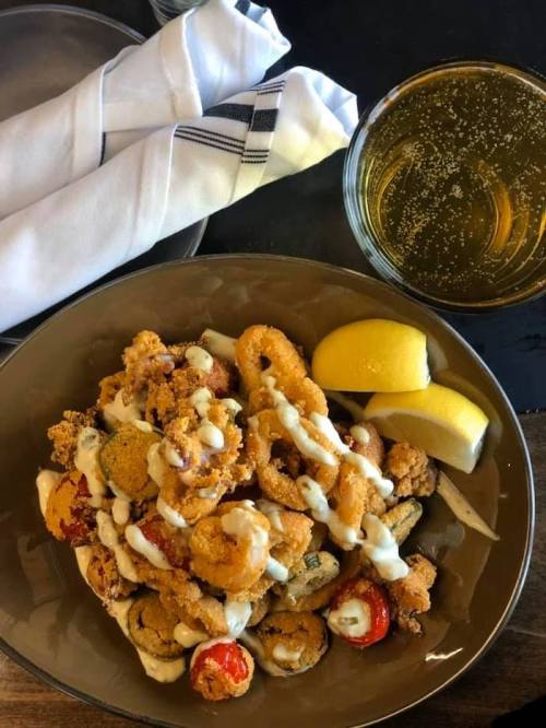 A bowl of fried seafood with sauce, garnished with lemon wedges, alongside a glass of sparkling drink and napkins.