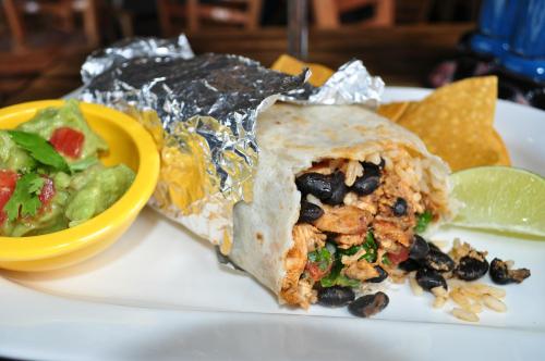 A close-up of a burrito wrapped in foil, served with chips, guacamole, and a lime wedge on a white plate.