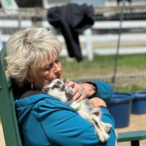 A woman with short blonde hair sits in a chair, holding a small goat in her arms, smiling in a sunny outdoor setting.