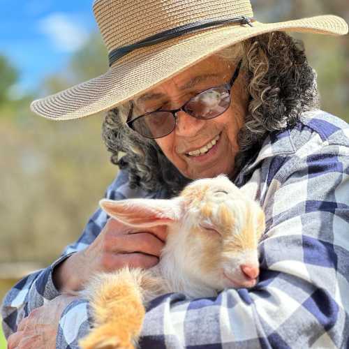 A smiling person in a hat holds a sleeping goat, surrounded by nature.