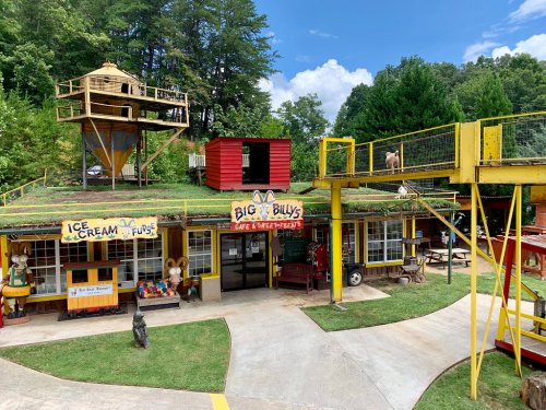 Colorful outdoor scene featuring a quirky café with a treehouse, red building, and playful decorations in a green setting.