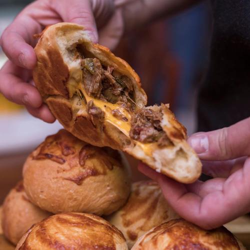 A person holds a cheesy, stuffed bread roll, revealing savory filling, with more rolls stacked in the background.