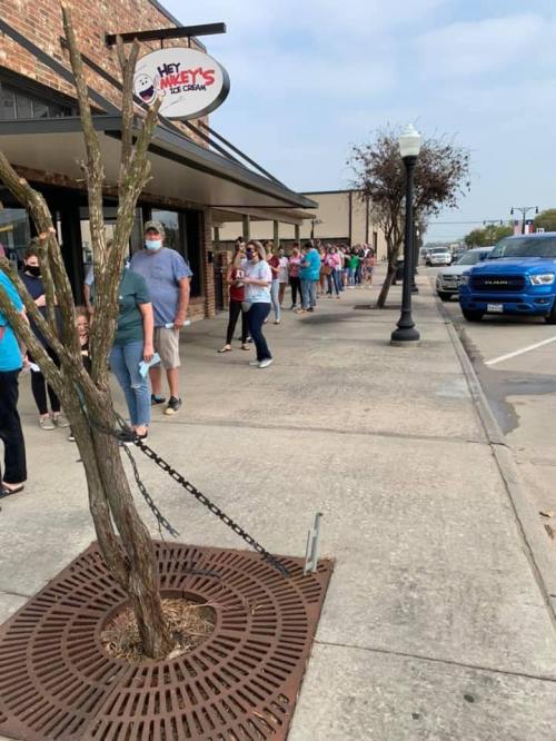 A long line of people wearing masks waits outside an ice cream shop on a sunny day.