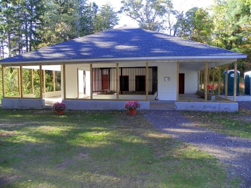 A single-story building with a porch, surrounded by trees, featuring a fenced area and potted flowers in front.