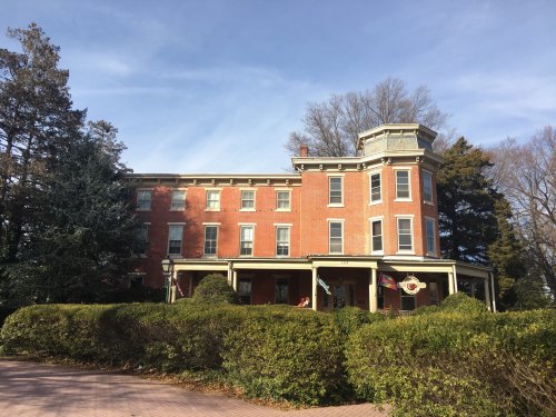 A large, historic red brick building with a wraparound porch, surrounded by greenery and trees under a clear blue sky.