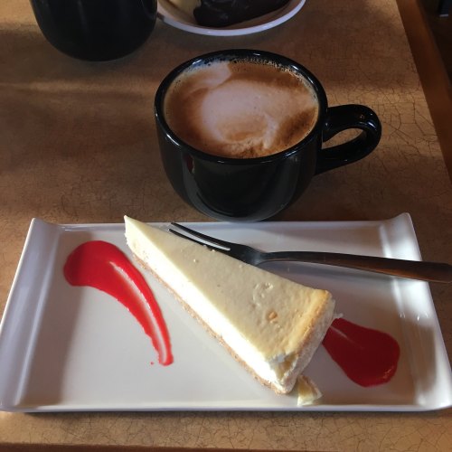 A slice of cheesecake on a plate with raspberry sauce, next to a black cup of cappuccino on a table.