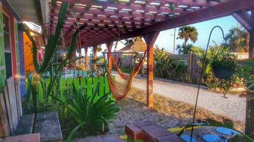 A colorful outdoor space with a hammock under a pergola, surrounded by plants and a sandy path leading to the beach.