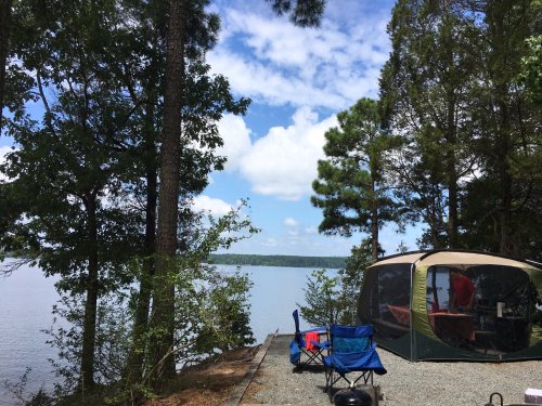 A serene lakeside campsite with a tent, chairs, and trees under a blue sky with fluffy clouds.