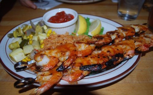 A plate of grilled shrimp skewers served with rice, vegetables, lemon wedges, and a small bowl of salsa.