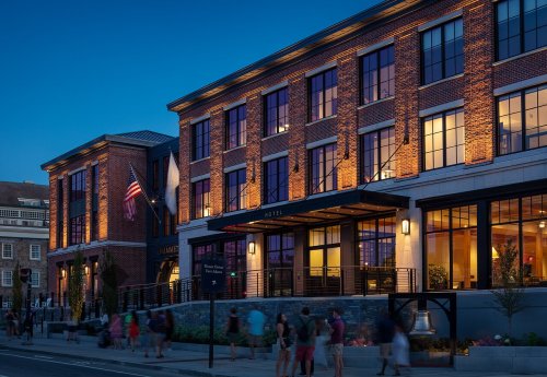 A modern hotel building with brick facade, illuminated at dusk, featuring large windows and people walking by.
