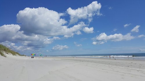 A serene beach scene with soft sand, gentle waves, colorful umbrellas, and fluffy clouds under a bright blue sky.