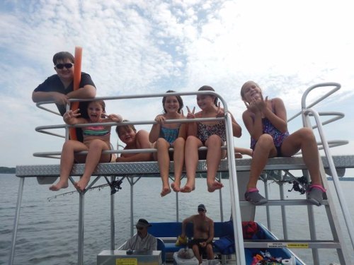 A group of five children in swimsuits sit on a boat, smiling and posing, with a man in the background.
