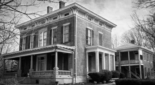 A historic brick house with columns, featuring a large porch and a neighboring building, set against a cloudy sky.