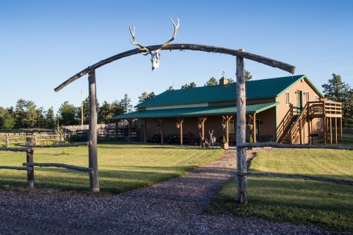 A rustic building with a green roof, framed by a wooden arch with deer antlers, set in a grassy landscape.