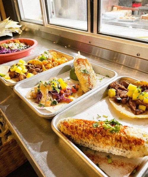 A variety of colorful tacos and dishes on metal trays, featuring vibrant toppings and garnishes.