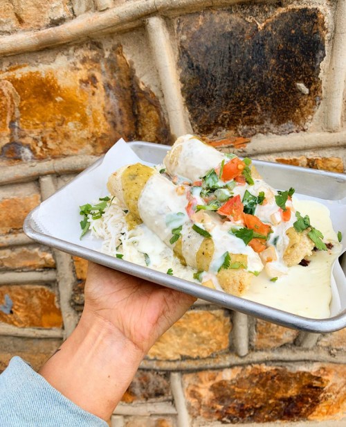 A hand holding a tray of rolled tacos topped with creamy sauce, diced tomatoes, and cilantro against a textured wall.