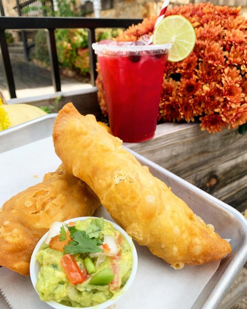 A plate with two golden empanadas and a small bowl of guacamole, alongside a colorful drink with a lime slice.
