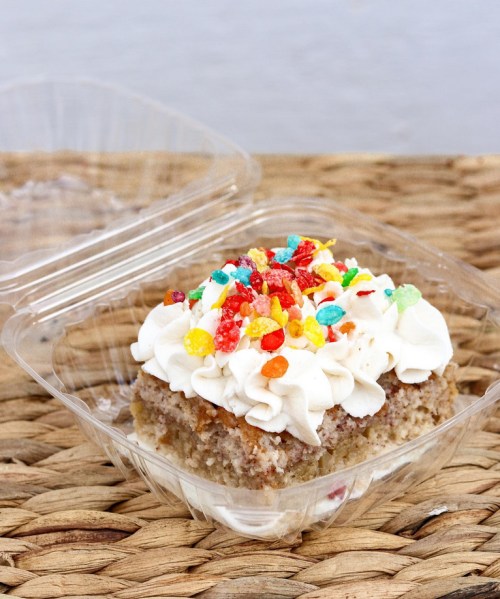 A dessert in a clear container, featuring a layered cake topped with whipped cream and colorful candy sprinkles.
